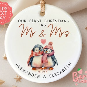 May include: White ceramic Christmas ornament with a penguin couple wearing scarves and hats. The ornament says "Our First Christmas as Mr & Mrs Alexander & Elizabeth 2023".