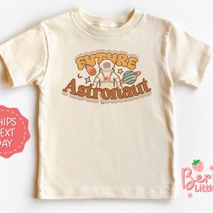 May include: A cream colored short-sleeved t-shirt with the text "FUTURE ASTRONAUT" in a retro font. The text is surrounded by a colorful design of a cartoon astronaut, a comet, a crescent moon, and a planet.