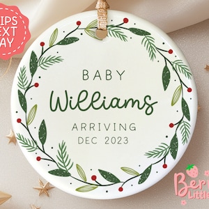 May include: White ceramic ornament with a green and red wreath design. The ornament says "Baby Williams Arriving Dec 2023".