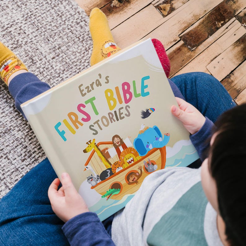 May include: A child is holding a colorful children's book titled "Ezra's First Bible Stories." The book cover features an illustration of Noah's Ark with various animals, including a giraffe, lion, and elephant. The child is wearing blue jeans and yellow socks.