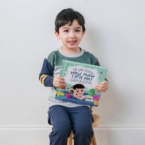 May include: A young boy is holding a children's book titled "Do you know how much I love you Grandpa". The book has a green cover with a cartoon illustration of a boy drawing.