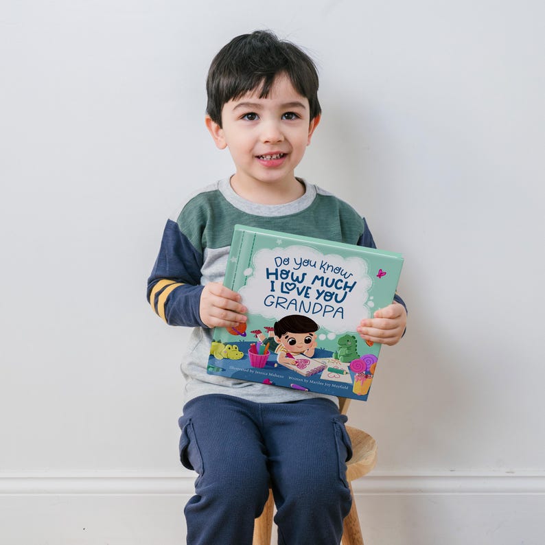 May include: A young boy is holding a children's book titled "Do you know how much I love you Grandpa". The book has a green cover with a cartoon illustration of a boy drawing.