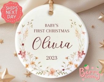 May include: A white ceramic bauble with a watercolour floral wreath design. The bauble reads "Baby's First Christmas Olivia 2023".