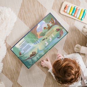 以下が含まれることがあります： 子供は森の風景の中に動物が描かれたカラフルなイラストの入った本を読んでいます。本には「自然の調和」というタイトルと、動物たちが一緒に遊んでいる様子が書かれた文章があります。子供はベージュのカーペットの上に座っており、近くに木製のシロフォンがあります。