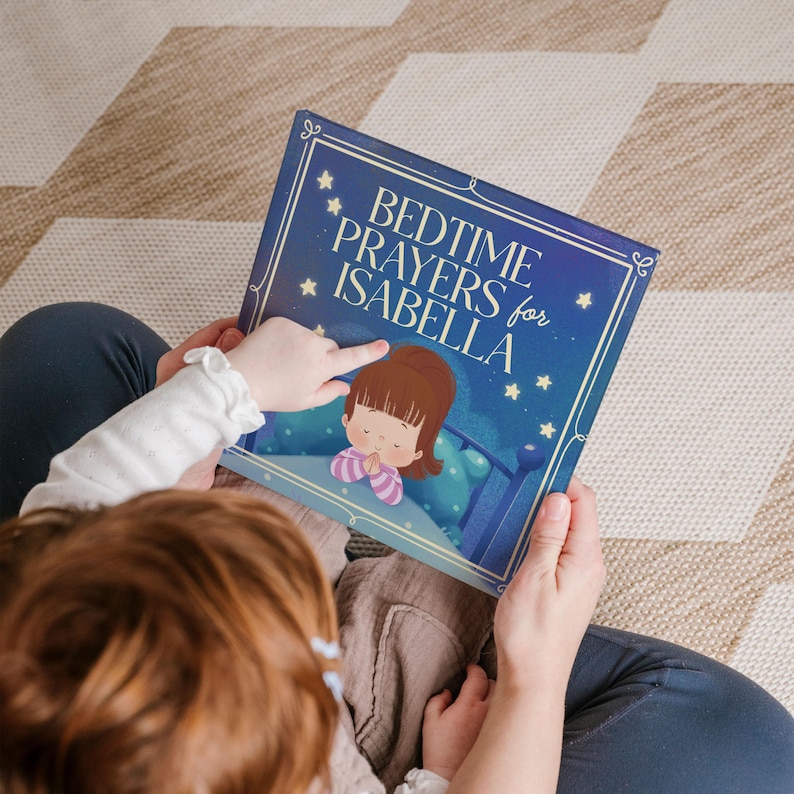 May include: A person holding a children's book titled "Bedtime Prayers for Isabella." The book cover is dark blue with white stars and a cartoon illustration of a child praying in bed. The background is a neutral-toned patterned rug.