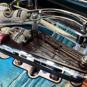 May include: A close-up of a pinball machine game featuring a wooden plank with two white scuba tanks and a rope. The plank is attached to a metal frame with a black rubber bumper.