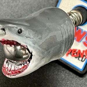 May include: A shark-themed bottle stopper with a gray and white painted shark head, open mouth revealing sharp teeth and a silver ball. The stopper has a spring and a black base with red and white text.