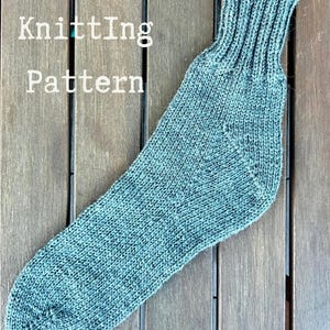 Puede incluir: Un calcetín tejido de color verde azulado con un puño acanalado. El calcetín se muestra sobre una superficie de madera. El texto "Knitting Pattern" es visible en letras blancas. El calcetín está hecho de un hilo texturizado.