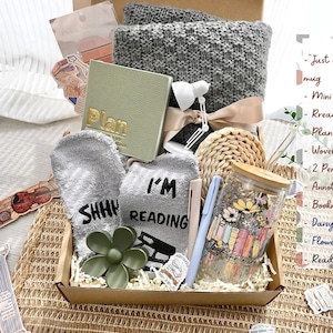 May include: A gift box filled with reading-themed items. Includes a gray knitted blanket, a green planner, reading socks, a glass mug with a floral design, pens, and a flower claw. The box is presented on a woven surface.