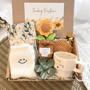 Pode incluir: Caixa de presente com uma caneca com a inscrição "hug in a mug", meias, tecido com estampa floral e um girassol de crochê. Há também um objeto verde em forma de flor. Apresentado em uma caixa marrom.