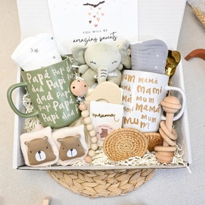 Op de afbeelding: Een cadeauverpakking voor baby's met een groene mok met de tekst "Papá Papi dad", een witte mok met de tekst "mum mamá", een pluche olifant, sokken, een fopspeen en een houten rammelaar. De doos bevat een kaart met "You will be amazing parents".