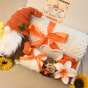 May include: A gift box filled with a gnome, a white cable-knit blanket, and orange accents. The gnome has an orange knit hat and a white beard. The box also contains sunflowers, a heart-shaped dish, and a card with ghosts.