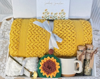 May include: A yellow knitted throw blanket in a gift box with a white ribbon. The box also contains two small glass bottles with cork tops, two white towels, a white mug, and a crocheted sunflower.