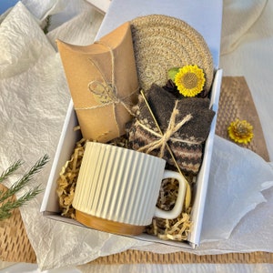 May include: A white mug with a ribbed design, a brown and white striped sock, a brown paper bag, a woven coaster, and a yellow flower are arranged in a white gift box with brown shredded paper.