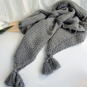 May include: A gray knitted throw blanket with tassels. The blanket is made of a soft, cozy material and has a textured pattern.