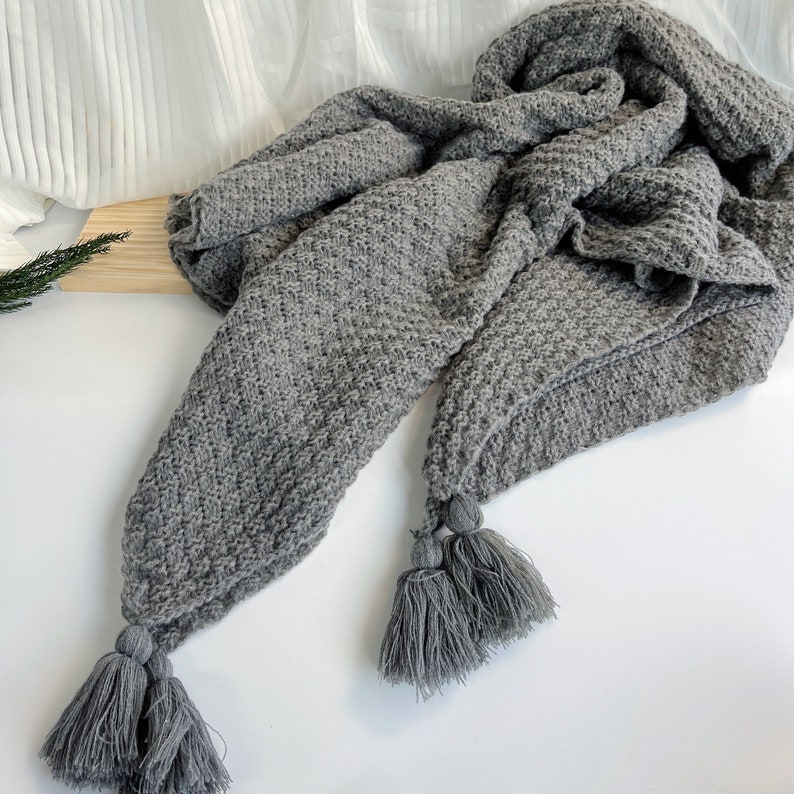 May include: A gray knitted throw blanket with tassels. The blanket is made of a soft, cozy material and has a textured pattern.