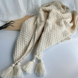 May include: A cream-colored knitted throw blanket with a diamond pattern and three tassels.