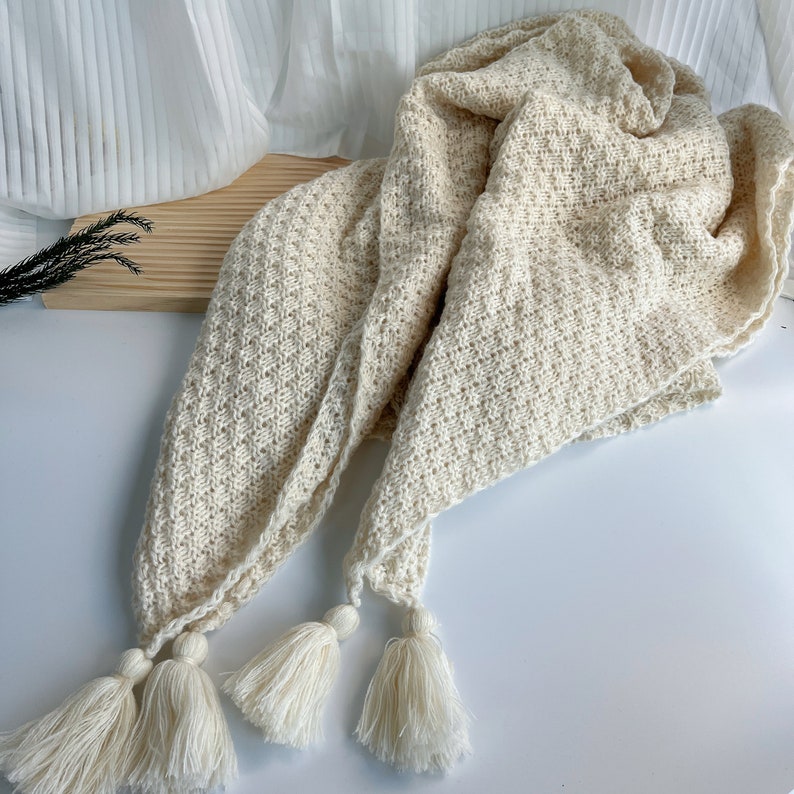 May include: A cream-colored knitted throw blanket with a diamond pattern and three tassels.