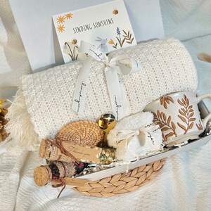 May include: A white knitted throw blanket, a white ceramic mug with brown leaf design, a small white towel, a small wicker basket, a small glass jar with a cork lid, a small brown wicker coaster, and a card that says "Sending Sunshine" are arranged in a wicker basket.
