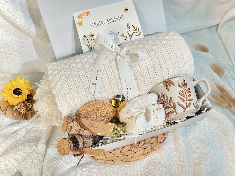 May include: A white knitted throw blanket, a white ceramic mug with brown leaf design, a small white towel, a small wicker basket, a small glass jar with a cork lid, a small brown wicker coaster, and a card that says "Sending Sunshine" are arranged in a wicker basket.