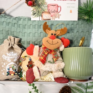 Pode incluir: Uma cesta de presente com tema natalino com uma caneca verde, um enfeite de rena, um saco de juta com o texto "Merry Christmas" e um cartão de felicitações. A cesta inclui um cobertor verde texturizado e pinhas.