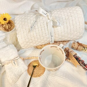 May include: A white knitted blanket with a white ribbon tied around it, a white mug with a brown floral design, a small white towel with a brown ribbon tied around it, and two small glass jars with brown lids and brown twine tied around them, filled with dried flowers and herbs. The jars have brown paper tags with the text "Thank you" written on them.