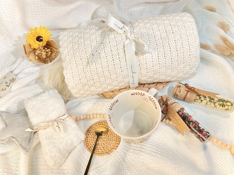 May include: A white knitted blanket with a white ribbon tied around it, a white mug with a brown floral design, a small white towel with a brown ribbon tied around it, and two small glass jars with brown lids and brown twine tied around them, filled with dried flowers and herbs. The jars have brown paper tags with the text "Thank you" written on them.