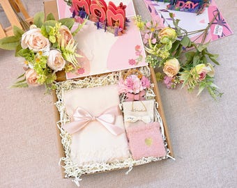 Op de afbeelding: Een cadeaudoos gevuld met een roze en witte deken, een klein roze zakje, een wit zakje met het woord "glimlachen" erop en een boeket roze bloemen. De doos is bekleed met bruine papieren strooisel.