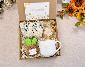 May include: A gift box filled with a white mug with daisy designs, a crocheted plant with a smiling face, a daisy-patterned fabric pouch, a crocheted coaster with a daisy, and a gold spoon. The box is lined with brown shredded paper and has a card that says "Sending Sunshine".