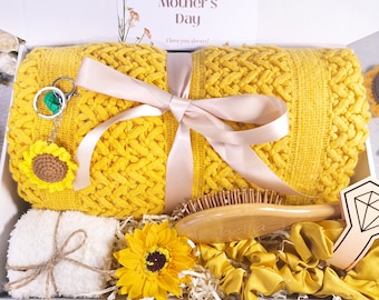 May include: A gift box filled with a yellow knitted blanket, a white towel, a wooden hairbrush, a sunflower-shaped keychain, a sunflower-shaped hair scrunchie, and a small wooden diamond-shaped ornament. The box is decorated with a pink ribbon and shredded paper.
