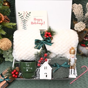 May include: A gift box containing a white textured throw blanket tied with a green ribbon, a dark green mug with the text "hug in a mug" and a heart, a white ceramic house, a grey folded textile, and a gold nutcracker ornament with red berries.