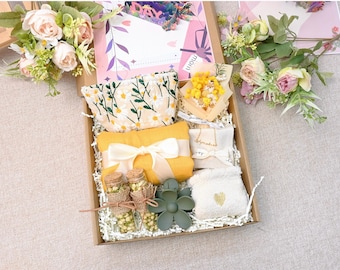 Op de afbeelding: Een cadeaudoos gevuld met verschillende items, waaronder een gele tas met bloemenprint, een gele sjaal, twee kleine glazen flesjes met gedroogde bloemen, een groen bloemachtig object, een witte washandje met een hartvormig ontwerp en een klein boeket gedroogde bloemen.