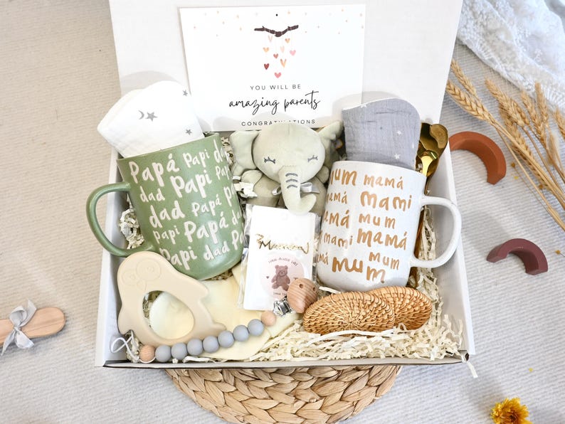 May include: A gift box filled with baby-themed items. Includes two mugs with "dad" and "mum" in various languages, a plush elephant, a teether, a card that says "You will be amazing parents", and other baby accessories.