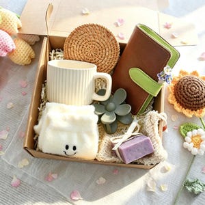 May include: A gift box containing a cream ribbed mug, a round woven coaster, a brown and lime green notebook, a gray flower-shaped hair clip, a pair of white socks with a smiley face, and a small purple soap bar tied with a white ribbon.