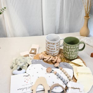 May include: A collection of baby items, including a plush elephant, bibs, a pacifier, and a wooden rattle, arranged on a woven mat. Two mugs are visible, one with "mum" and the other with "dad" printed on them. A card reads "You will be amazing parents."