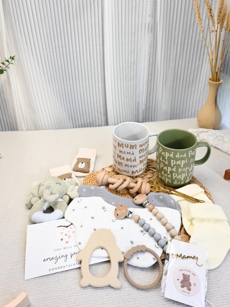 May include: A collection of baby items, including a plush elephant, bibs, a pacifier, and a wooden rattle, arranged on a woven mat. Two mugs are visible, one with "mum" and the other with "dad" printed on them. A card reads "You will be amazing parents."