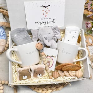 May include: A white gift box filled with baby items, including two white mugs with "mama" and "bear" printed on them. The box also contains a gray elephant plush, baby socks, and a card that reads "You will be amazing parents."