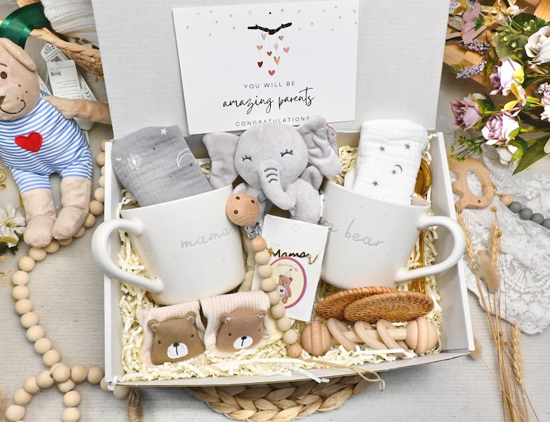 May include: A white gift box filled with baby items, including two white mugs with "mama" and "bear" printed on them. The box also contains a gray elephant plush, baby socks, and a card that reads "You will be amazing parents."