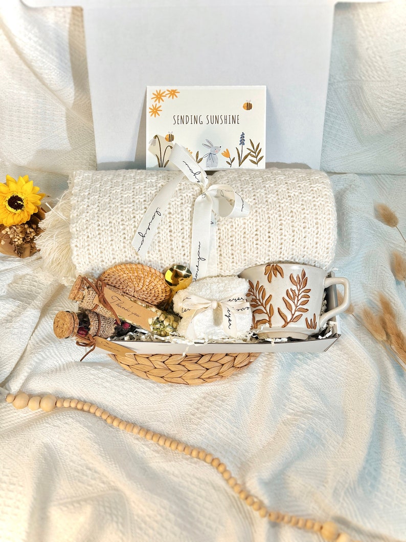 May include: A white wicker basket filled with a white knitted blanket, a white mug with brown leaf design, a small bottle of bath salts, a small white towel, a small bag of dried flowers, and a gold ornament. The basket is tied with a white ribbon with the words "Sending Sunshine" printed on it. The basket is sitting on a white surface with a wooden bead garland.