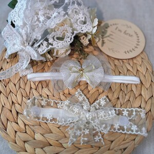 May include: Two white lace headbands with bows and a wooden sign that says "I'm here". The headbands and sign are on a woven straw mat.