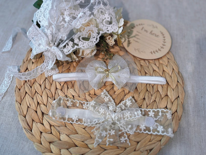 May include: Two white lace headbands with bows and a wooden sign that says "I'm here". The headbands and sign are on a woven straw mat.