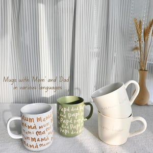 May include: A collection of ceramic mugs with various text designs. One mug is white with "mum mama" in gold lettering. Another is green with "Pap&aacute; dad Papi" in white. Two stacked cream-colored mugs say "mama bear" and "papa bear".