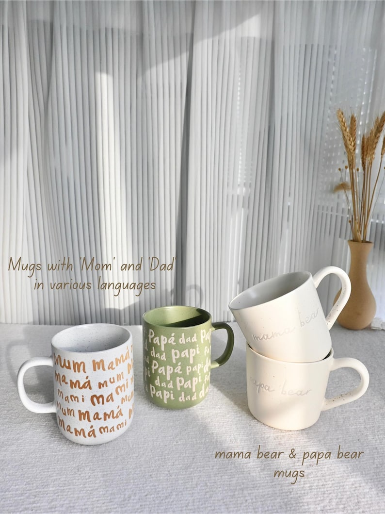 May include: A collection of ceramic mugs with various text designs. One mug is white with "mum mama" in gold lettering. Another is green with "Pap&aacute; dad Papi" in white. Two stacked cream-colored mugs say "mama bear" and "papa bear".