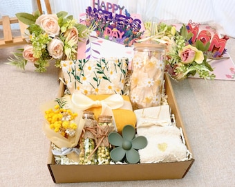 Op de afbeelding: Een bruine cadeaudoos gevuld met verschillende items, waaronder een gele handdoek, een bloemenpouch, een glazen pot met witte bloemen, een klein boeket gele bloemen, twee kleine glazen flesjes met gedroogde bloemen, een groene bloemvormige zeepbak en een witte handdoek met een hartvormig ontwerp. De doos is versierd met wit tissuepapier en een klein kaartje met de tekst "Happy Mother's Day".