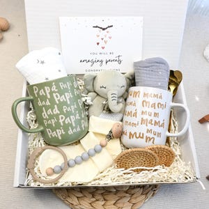 May include: A gift box for new parents, featuring two mugs with "Dad" and "Mom" inscripted, a plush elephant, baby accessories, and a card that reads "You will be amazing parents". The box is filled with shredded paper.