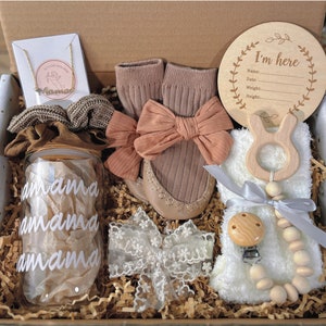 May include: A gift box filled with baby items, including a wooden sign that says "I'm here", a glass jar with the word "amama" written on it, a pair of brown socks, a wooden teether, a wooden bead necklace, and a white lace headband.