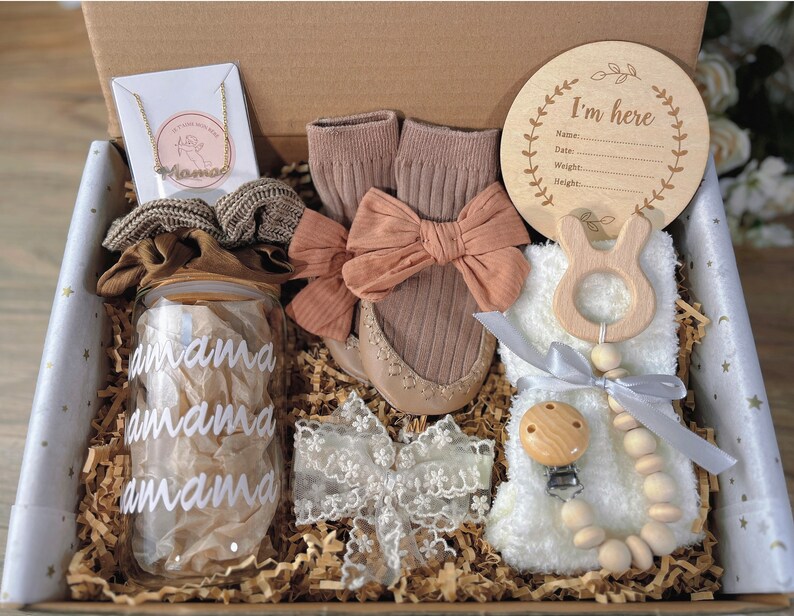 May include: A gift box filled with baby items, including a wooden sign that says "I'm here", a glass jar with the word "amama" written on it, a pair of brown socks, a wooden teether, a wooden bead necklace, and a white lace headband.