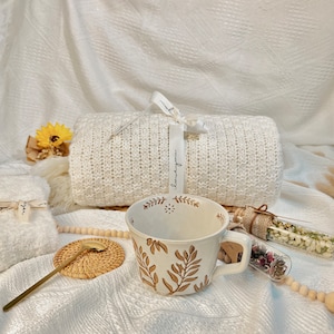 May include: A white ceramic mug with a brown floral design. The mug is sitting on a white knitted blanket with a wooden coaster and a gold spoon. There are two glass vials with dried flowers and a white knitted blanket in the background.