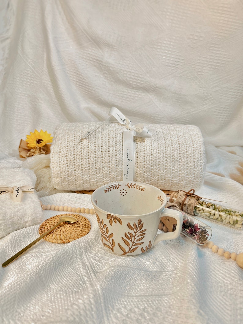 May include: A white ceramic mug with a brown floral design. The mug is sitting on a white knitted blanket with a wooden coaster and a gold spoon. There are two glass vials with dried flowers and a white knitted blanket in the background.