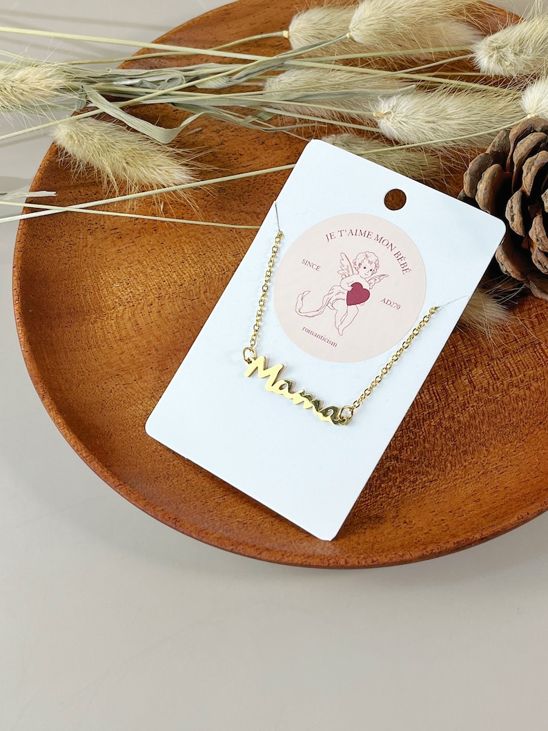 May include: A gold necklace with a cursive "Mama" pendant on a white card with the text "Je t'aime mon b&eacute;b&eacute; since AD 279" and a small illustration of a cherub with a heart.